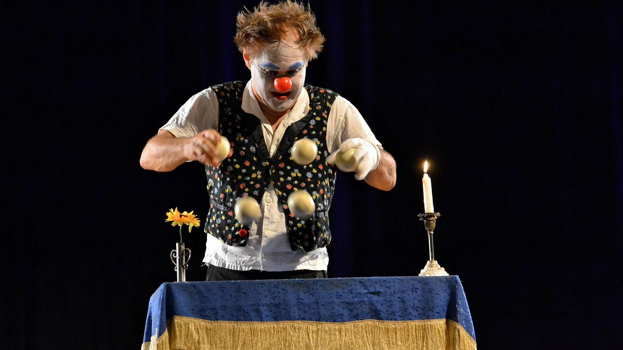 Calvero der Clown steht an einem kleinen Tisch mit einer Kerze und einer Blumenvase und jongliert mit vier Bällen.