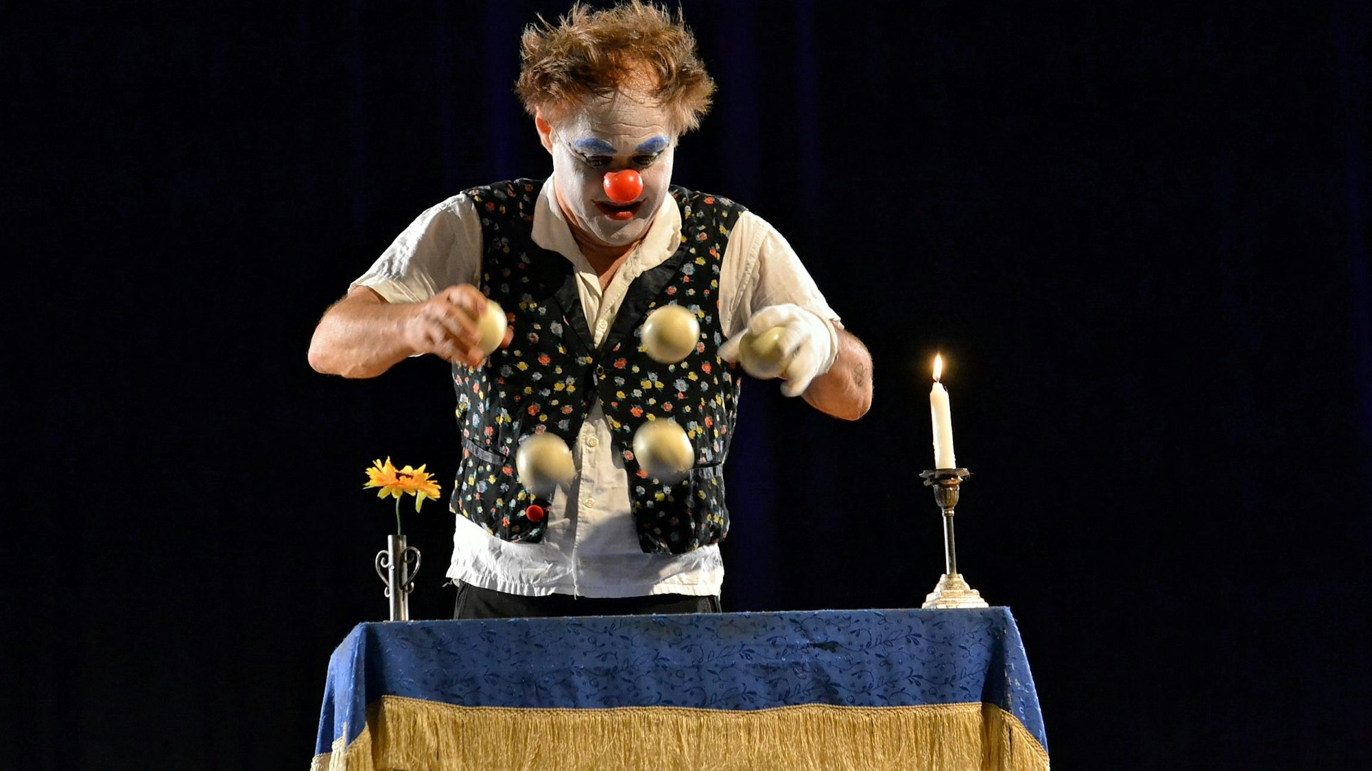 Calvero der Clown steht an einem kleinen Tisch mit einer Kerze und einer Blumenvase und jongliert mit vier Bällen.