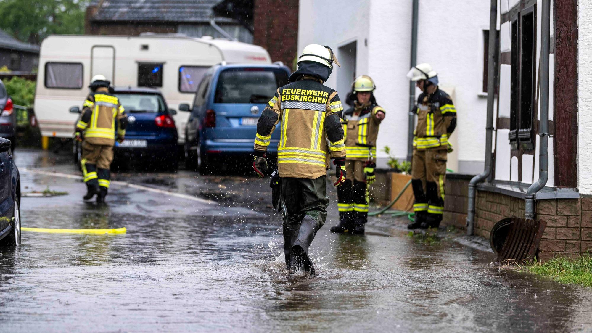 Feuerwehrleute stehen an einer Straße mit Häusern, auf der Wasser fließt.