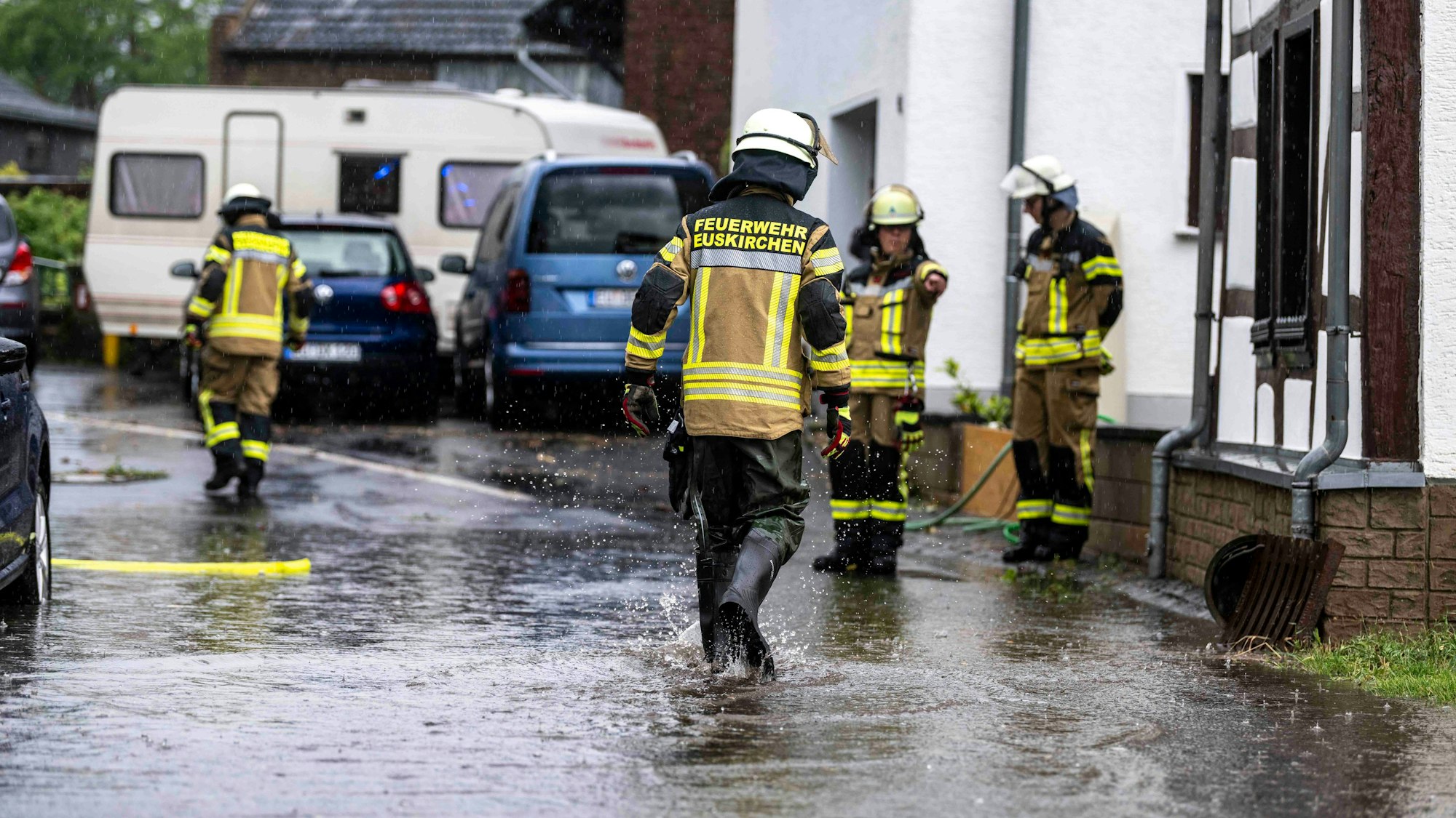 Feuerwehrleute stehen an einer Straße mit Häusern, auf der Wasser fließt.