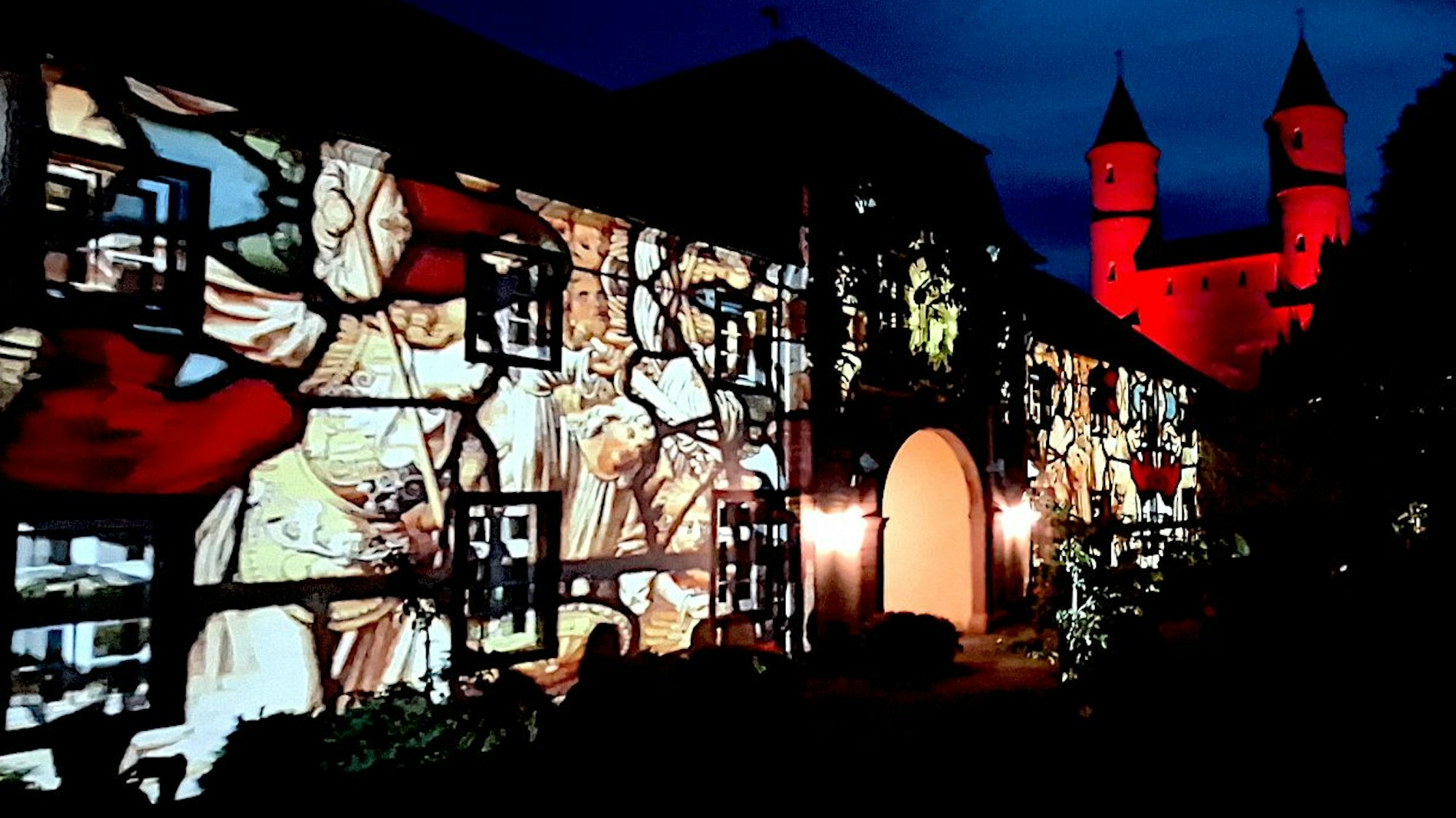 Auf dem langgestreckten Torbau ist eine biblische Szene in leuchtenden Farben zu sehen. Im Hintergrund werden die Zwillingstürme des Klosters rot vor dem nächtlichen Himmel angestrahlt.