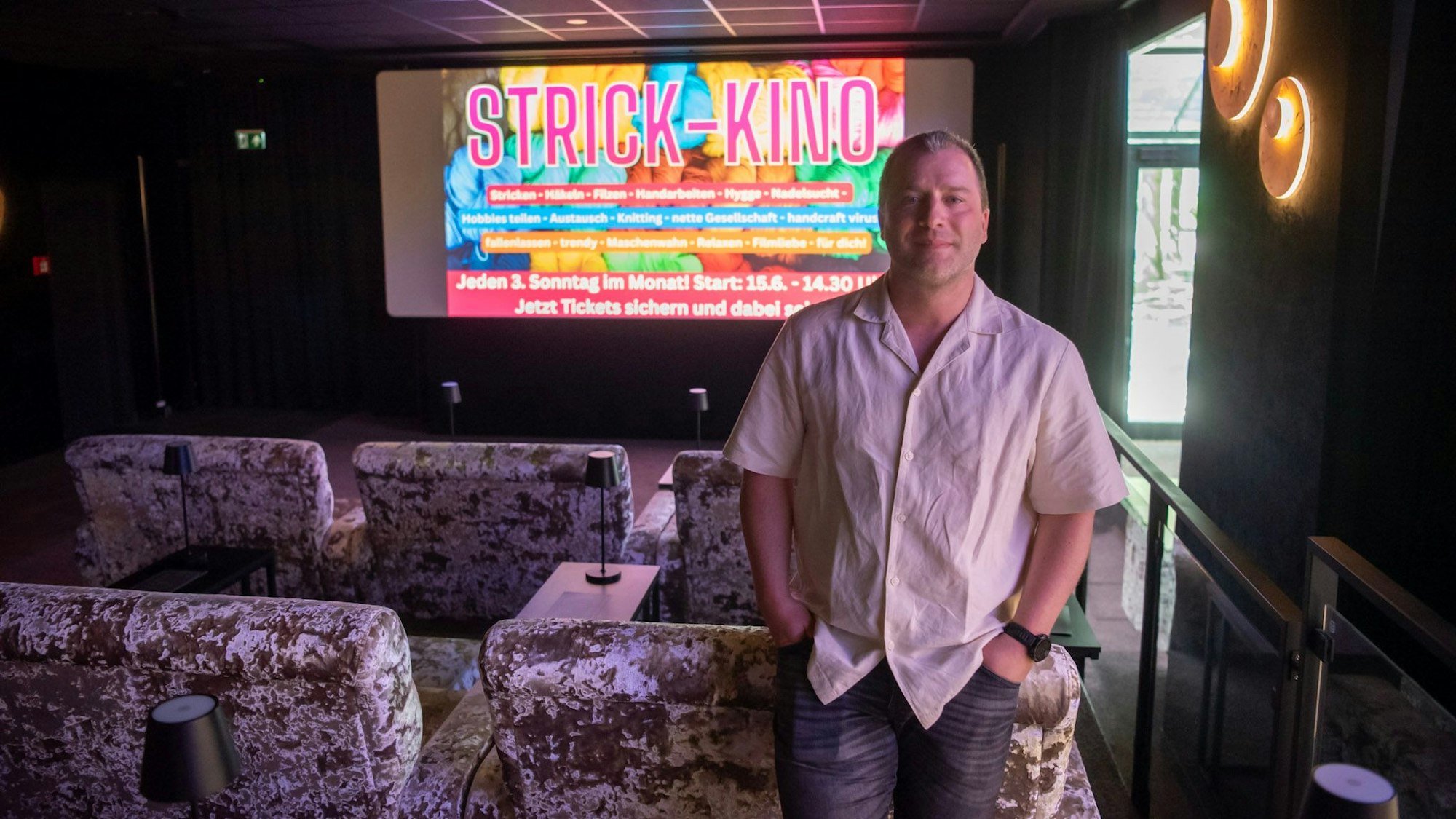 Sebastian Arenz steht an ein Plüschsofa gelehnt vor der Leinwand der Lichtspiellounge in Euskirchen, auf der in großen Buchstaben „Strick Kino“ steht.