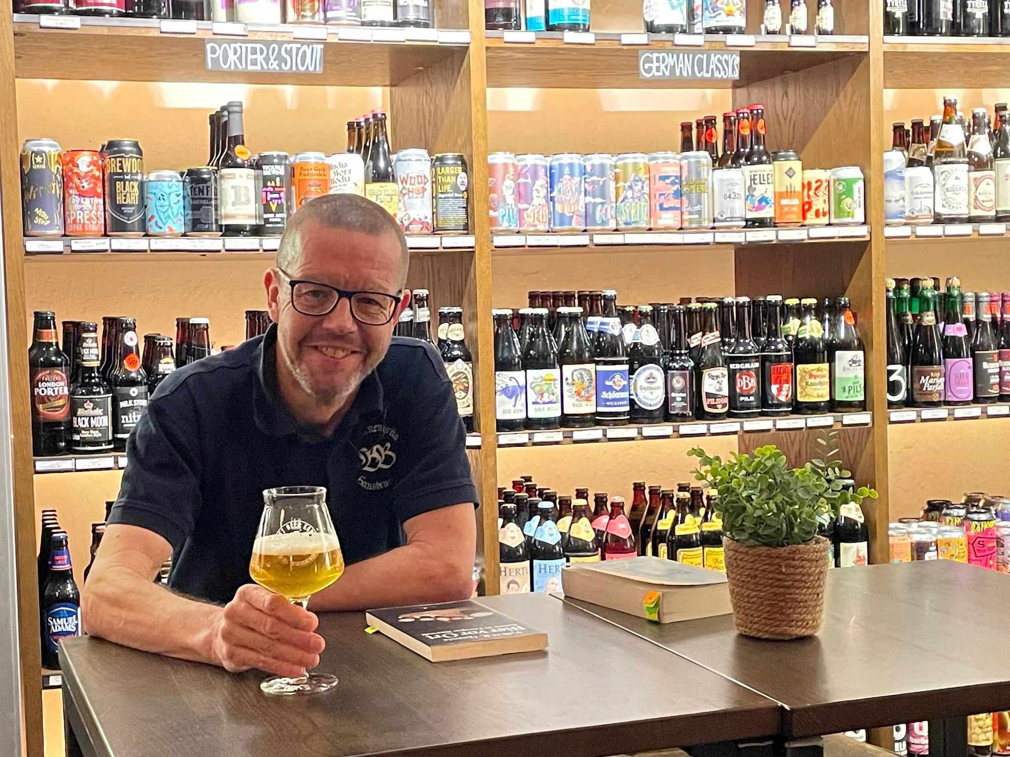 Auf dem Bild ist ein Mann mit Bierglas vor einem Regal mit vielen verschiedenen Biermarken zu sehen.