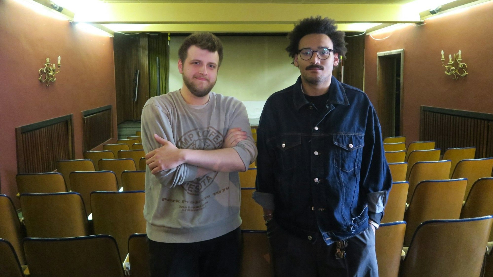 Zwei junge Männer stehen in einem altmodisch-anmutenden Kino-Saal.