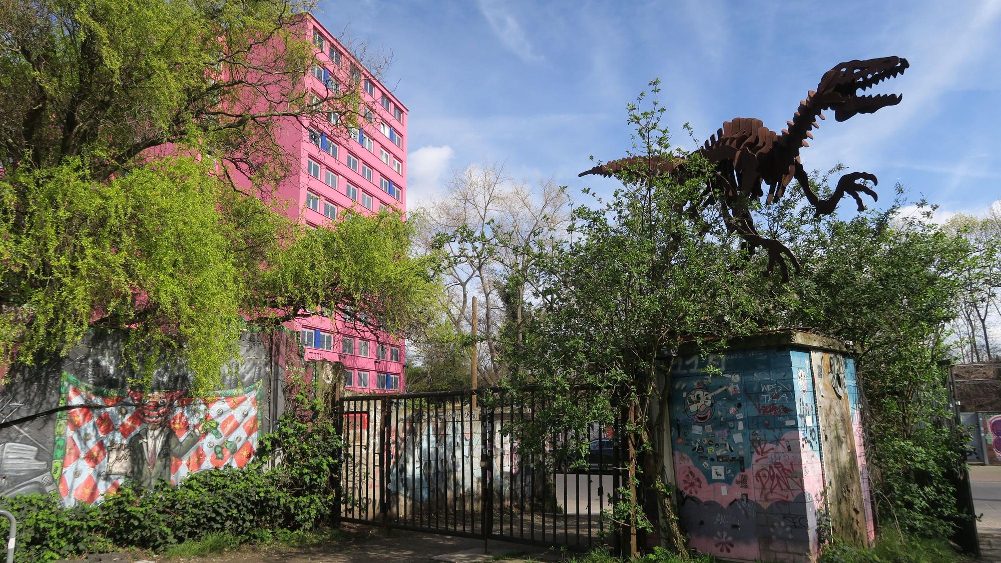 Hinter einem rostigen, halb zugewucherten Tor steht eine riesige Dinosaurier-Skulptur, auch ein mehrstöckiges pinkes Gebäude ist zu sehen.