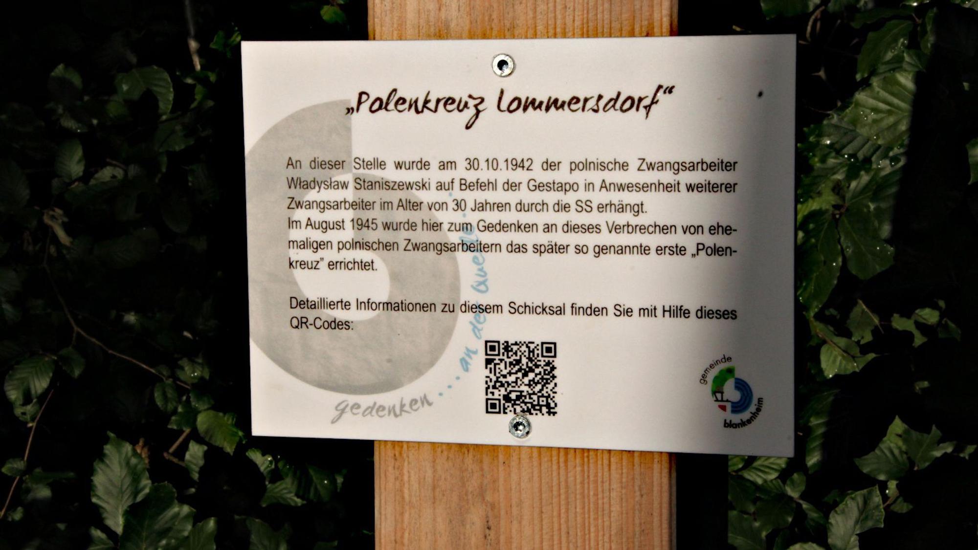 Ein Blatt mit der Überschrift Polenkreuz Lommersdorf und einem QR-Code hängt an einem Holzstab.