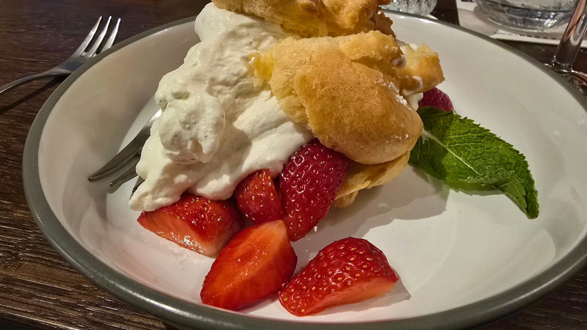 Das Dessert ist auf einem weißen Teller zu sehen: Windbeutel, aus dem die Creme quillt, daneben liegen Erdbeeren und ein Minzblatt.