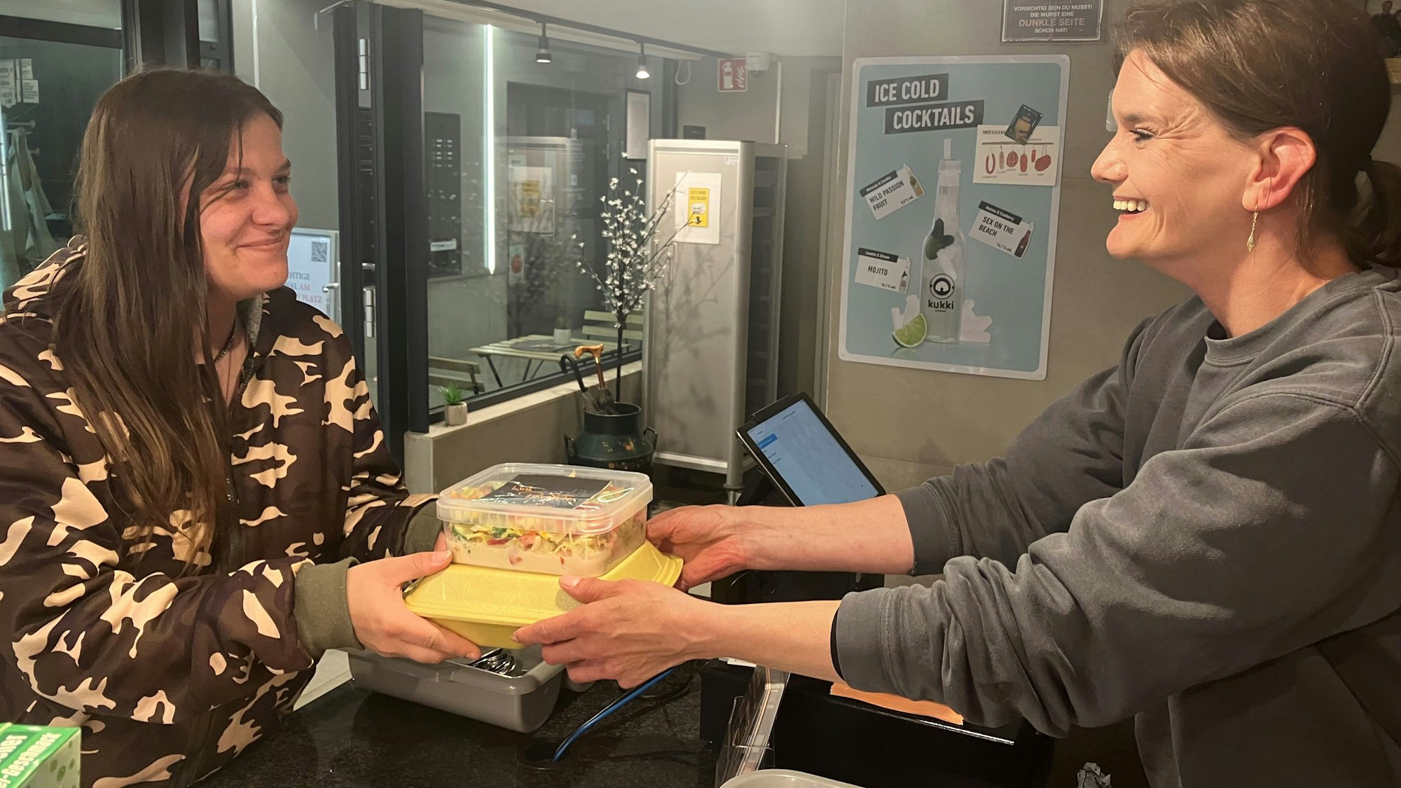 Blick auf zwei Frauen, die zwei Imbissschachteln über eine Theke reichen.