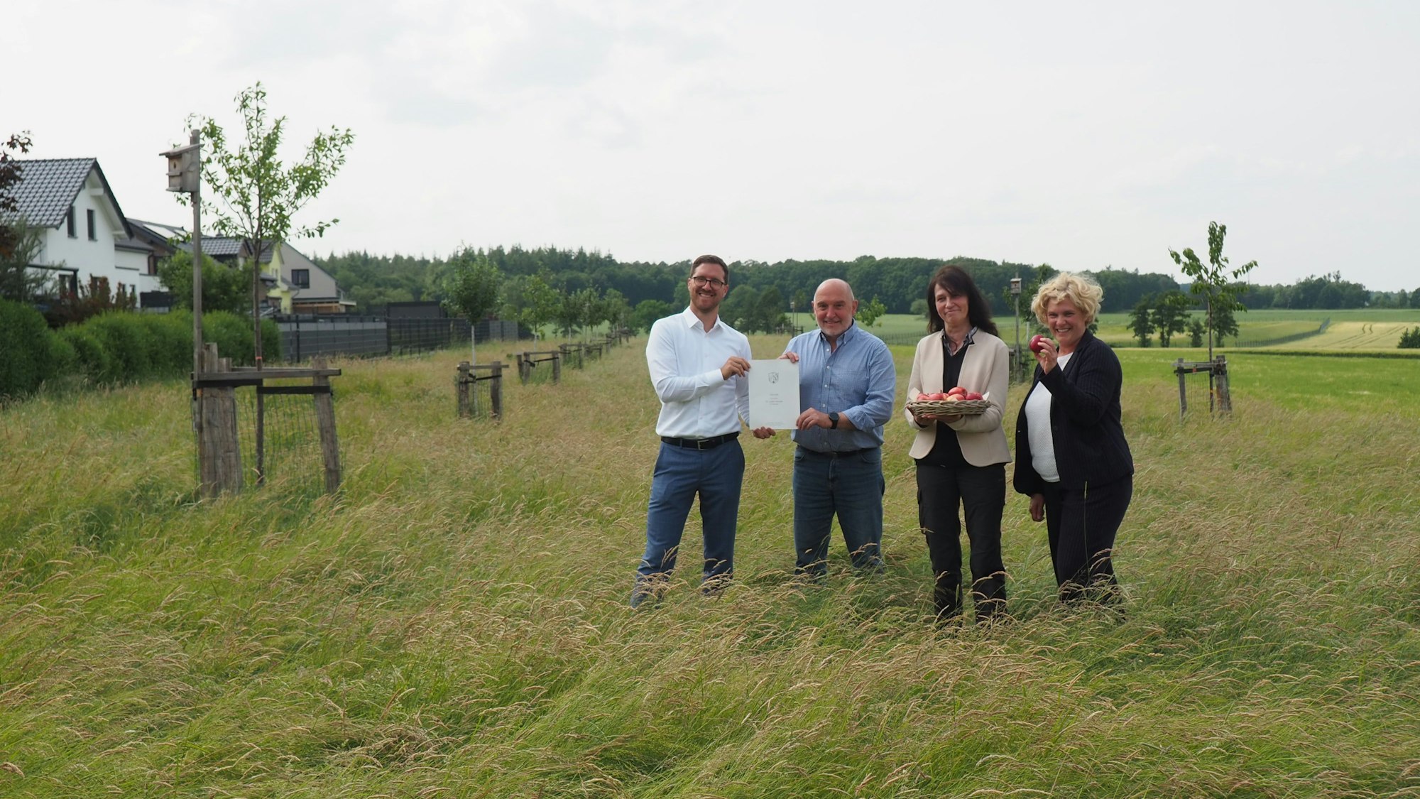 Zwei Männer und zwei Frauen stehen auf einer ungemähten Wiese zwischen jungen Obstbäumen.