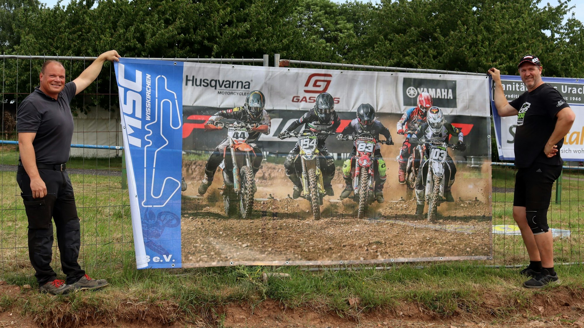 Herbert Maubach und Jens Hoch halten ein Werbeplakat in die Höhe, das sie an einer Absperrung befestigen wollen.