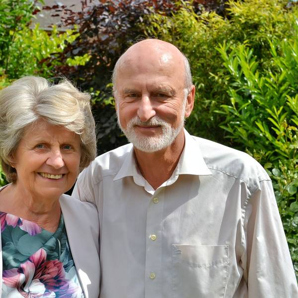 Annelies und Wolfram Göbel stehen vor Blätterwerk. Die älteren Menschen tragen ein Sakko mit einem Oberteil in Blumenmuster und ein Hemd.