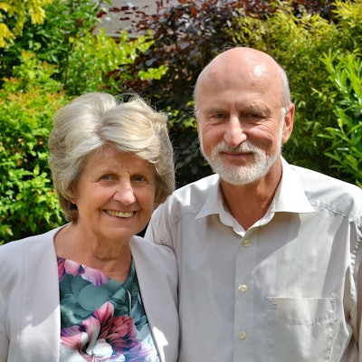 Annelies und Wolfram Göbel stehen vor Blätterwerk. Die älteren Menschen tragen ein Sakko mit einem Oberteil in Blumenmuster und ein Hemd.