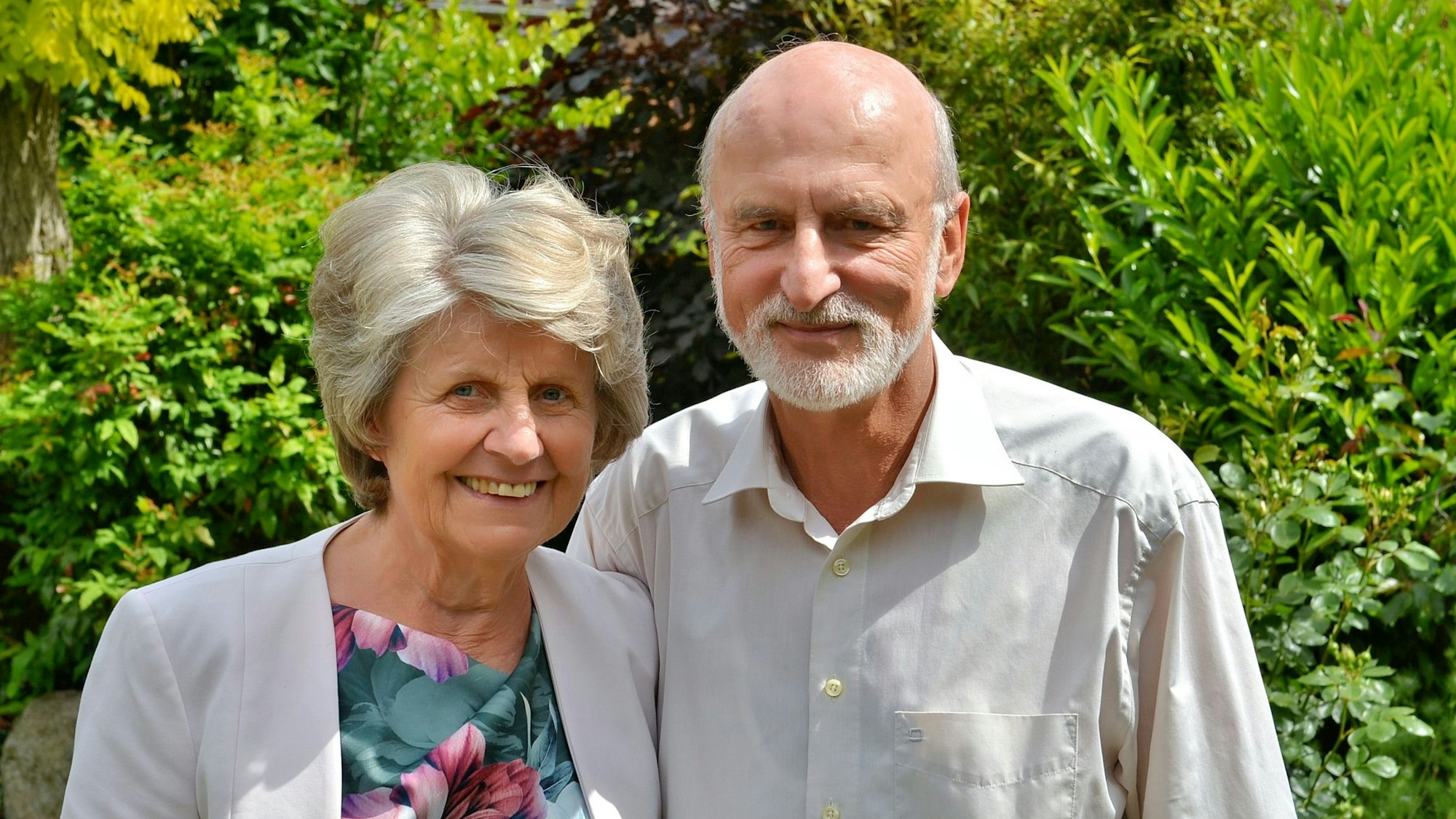 Annelies und Wolfram Göbel stehen vor Blätterwerk. Die älteren Menschen tragen ein Sakko mit einem Oberteil in Blumenmuster und ein Hemd.