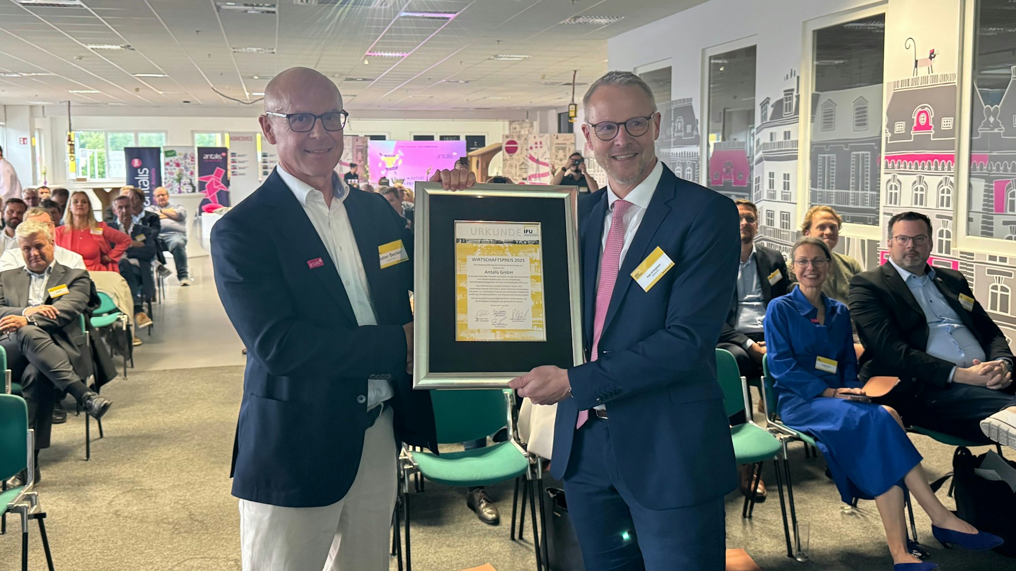 Auf dem Bild sind zwei Männer in Jackets bei der Übergabe einer Urkunde zu sehen.