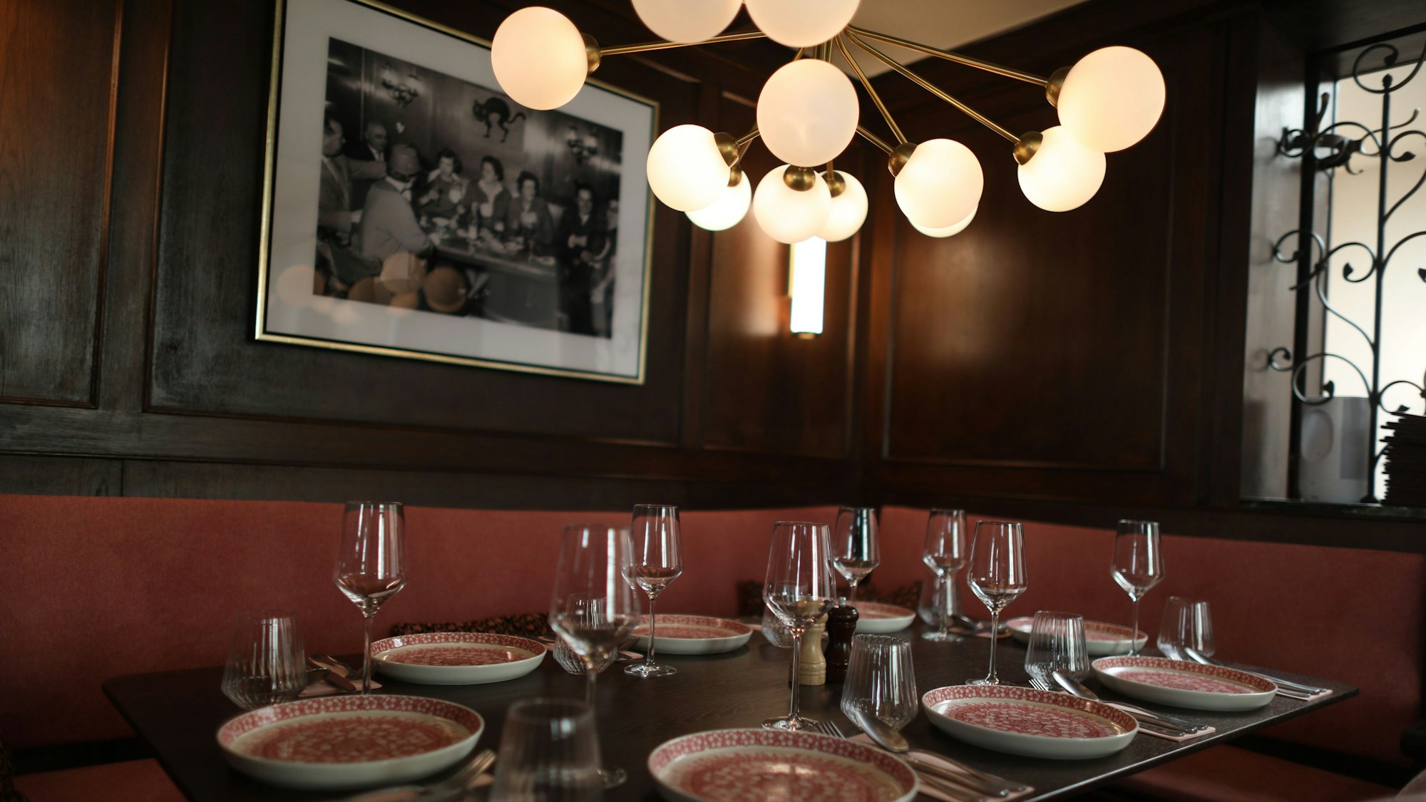 Ein eingedeckter Tisch unter einer mehrleuchtigen Lampe ist zu sehen. Über der Sitzbank auf der holzvertäfelten Wand ein Schwarzweißfoto in Rahmen.