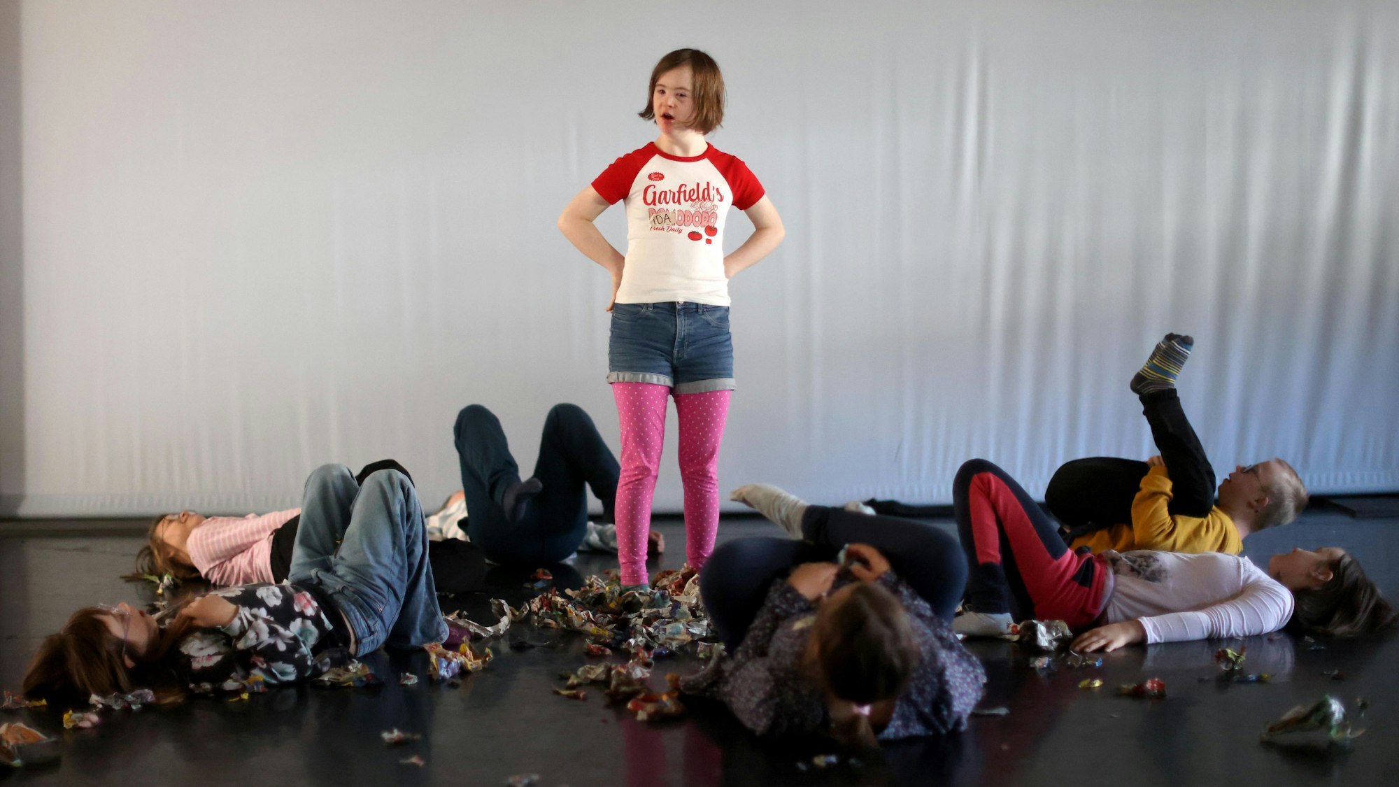 Ida hat die Hände in die Hüften gestemmt, die anderen Darstellenden sind rücklings umgefallen und liegen zwischen den bunten Papierkugeln auf dem schwarzen Boden.