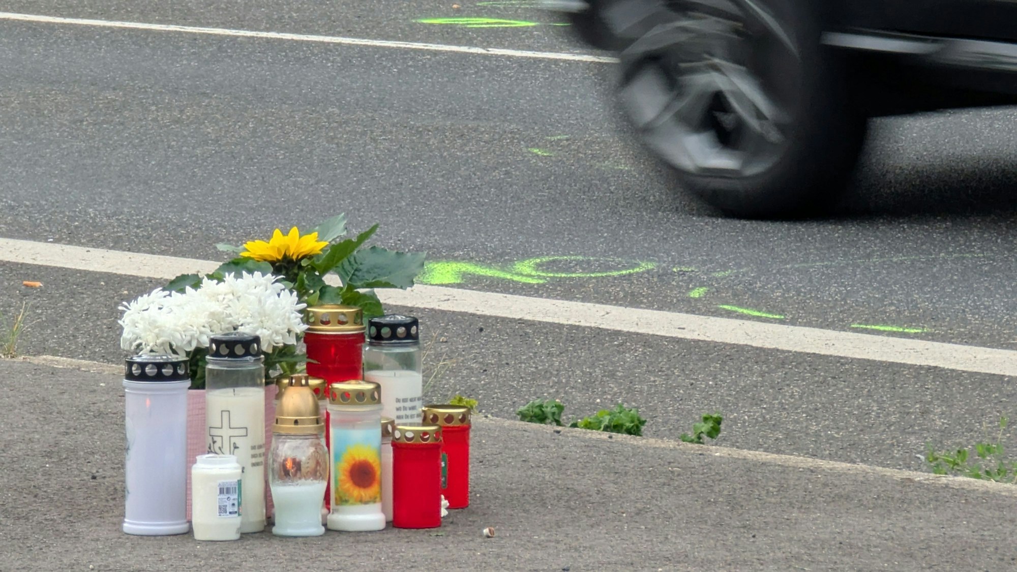 Das Foto zeigt Blumen und Kerzen am Straßenrand. Auf der Fahrbahn sind noch die Markierungen der Unfallermittler zu sehen.