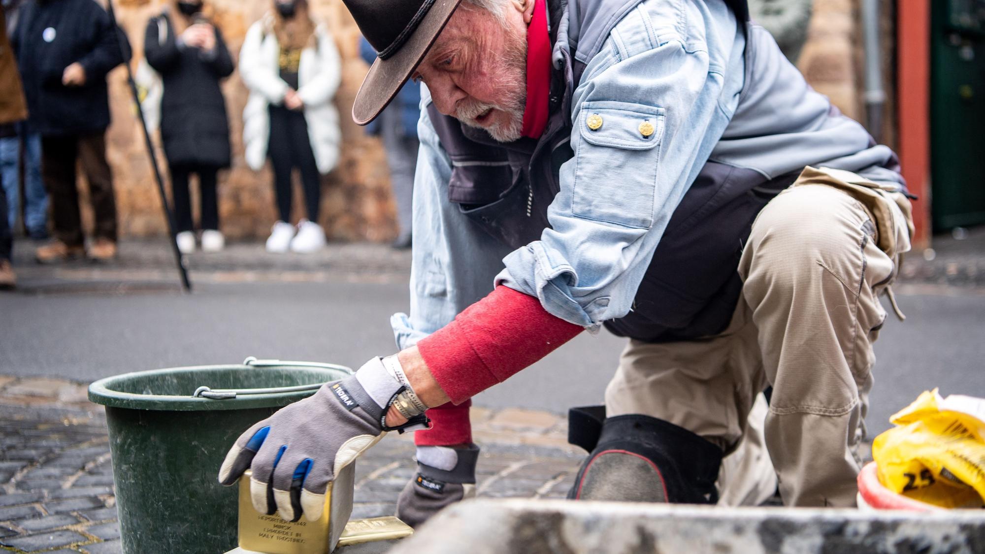 Künstler Gunter Demnig, ein älterer Herr mit Hut, hat einen Stolperstein gegriffen. Er trägt Handschuhe und kniet auf Pflastersteinen.