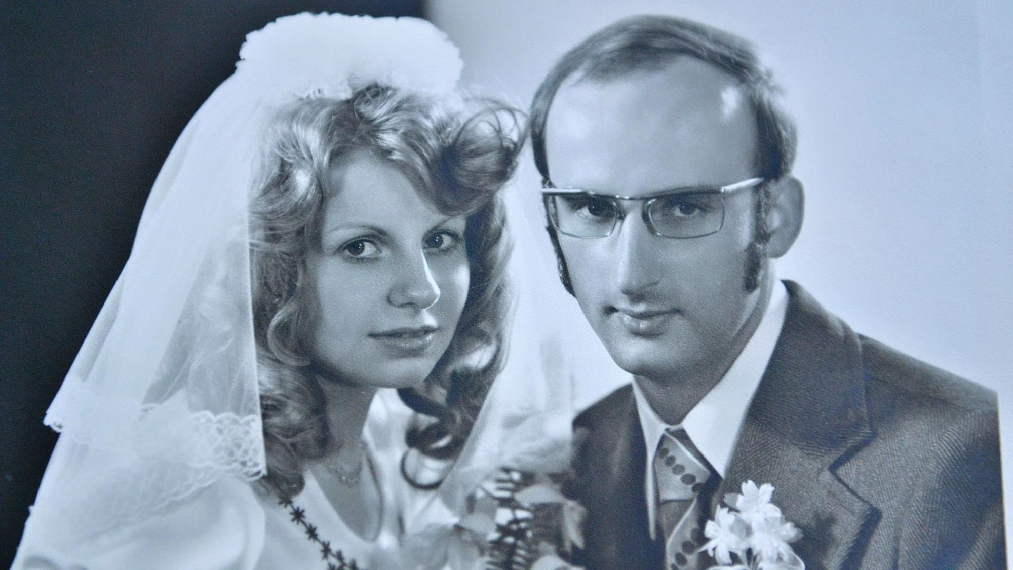 Die Eheleute Annelies und Wolfram Göbel posieren in Hochzeitskleid und Anzug auf einem Foto in Schwarz-weiß. Sie hat welliges Haar. Erträgt eine Brille und Koteletten.