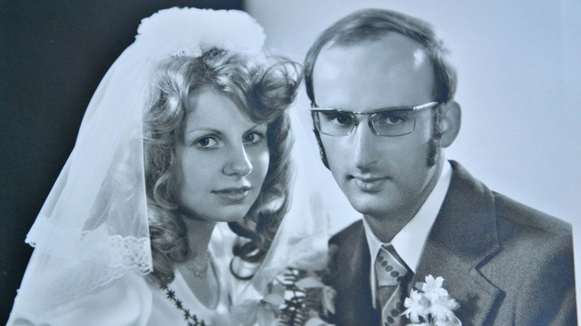 Die Eheleute Annelies und Wolfram Göbel posieren in Hochzeitskleid und Anzug auf einem Foto in Schwarz-weiß. Sie hat welliges Haar. Erträgt eine Brille und Koteletten.