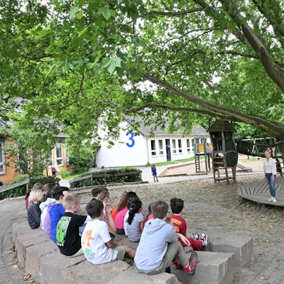 Auf Steinblöcken sitzen Kinder unter dem Blätterdach einer Platane.