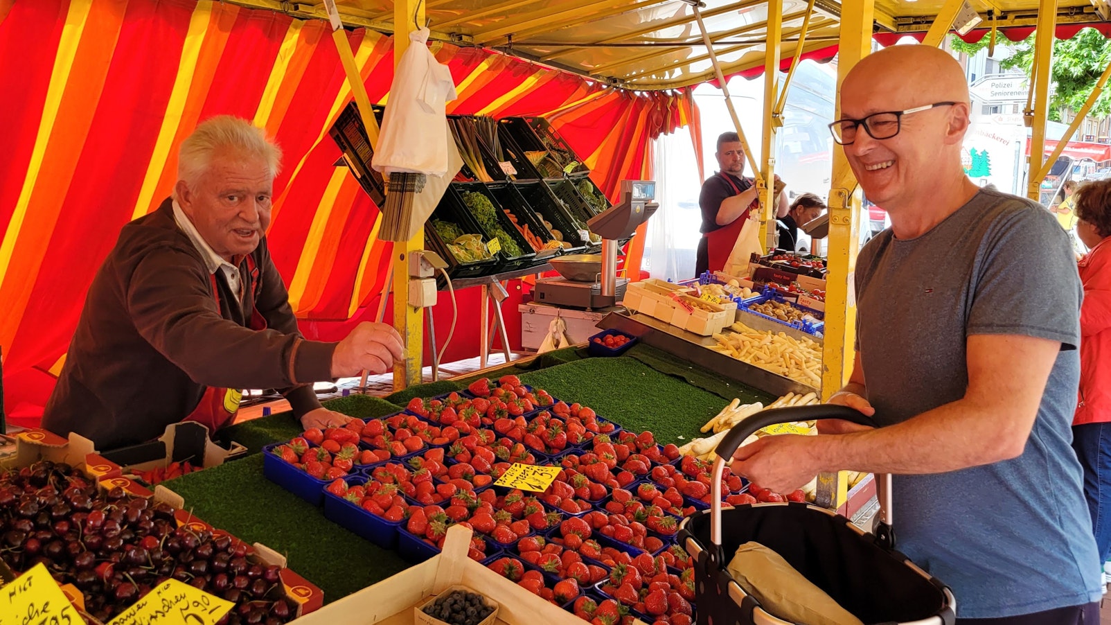 Ein Mann bedient einen Kunden auf einem Wochenmarkt, der gerade Erdbeeren gekauft hat.