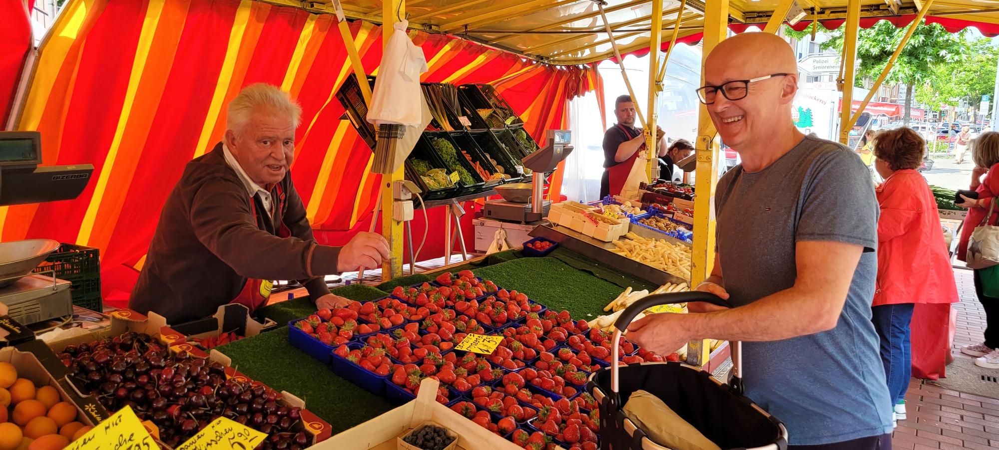 Auf dem Bild ist ein Obst- und Gemüsestand mit einem Händler und einem Kunden zu sehen.