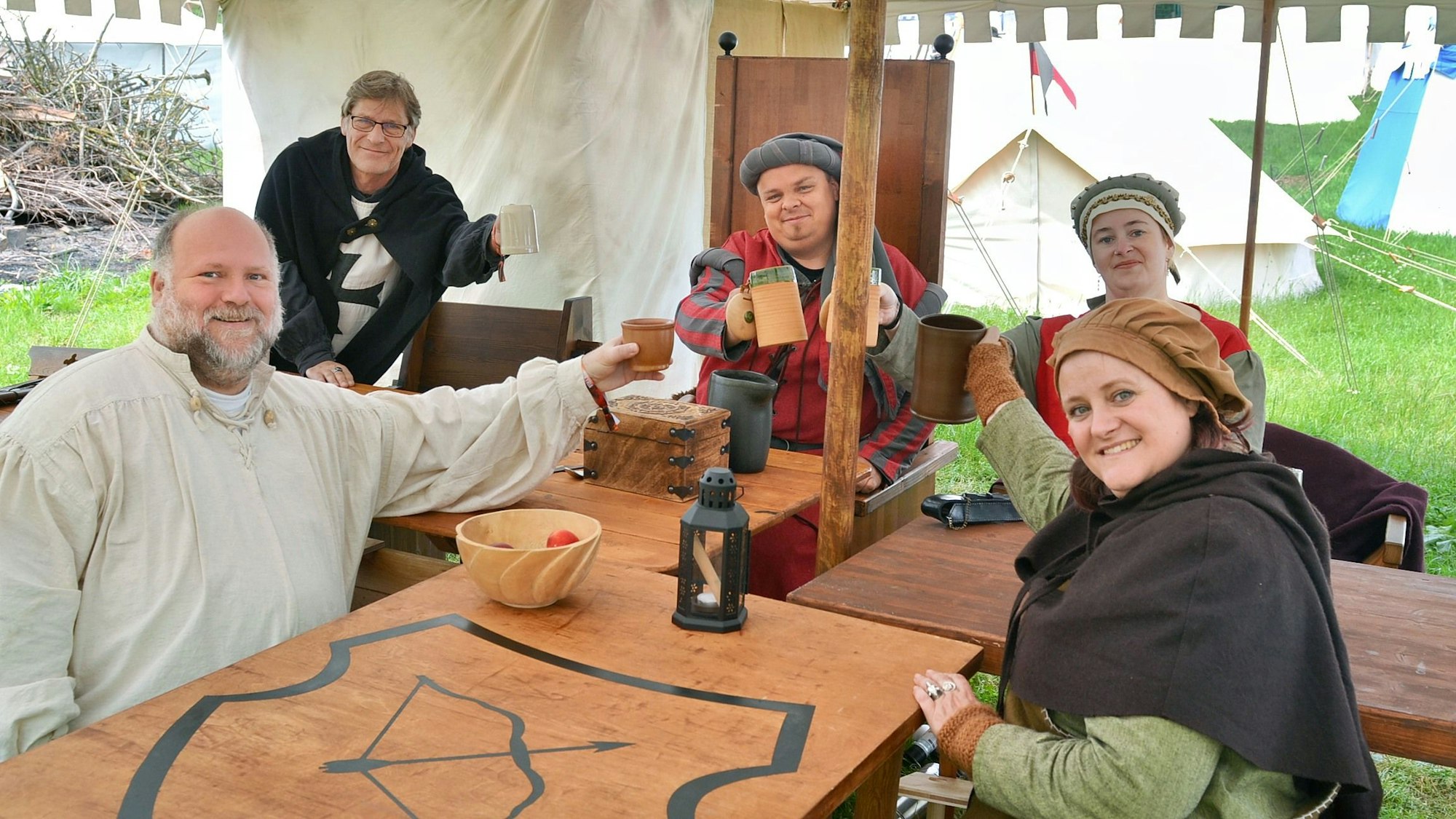 Eine Gruppe mittelalterlich gekleideter Leute sitzt an einem Tisch und prostet sich mit Bierkrügen zu.