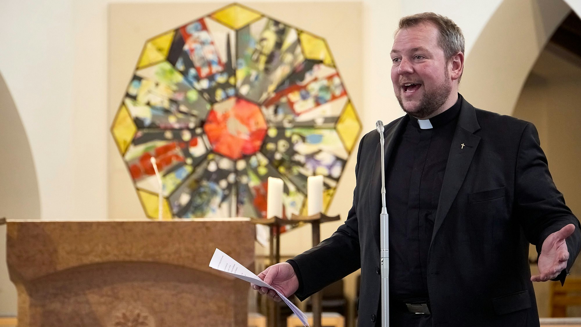 Pfarrer Thomas Schlütter steht in Schwarz gekleidet an einem Mikrofon und hält ein Blatt Papier in der Hand. Seine Arme sind ausgestreckt, sein Gesichtsausdruck ist enthusiastisch. Im Hintergrund ist ein Gemälde eines oktogonalen Kirchenfensters zu sehen.