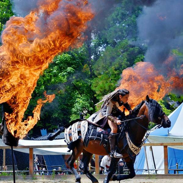 Eine Frau mit geflochtenen Haaren und Kampfausrüstung reitet zwischen zwei Feuertonnen hindurch.