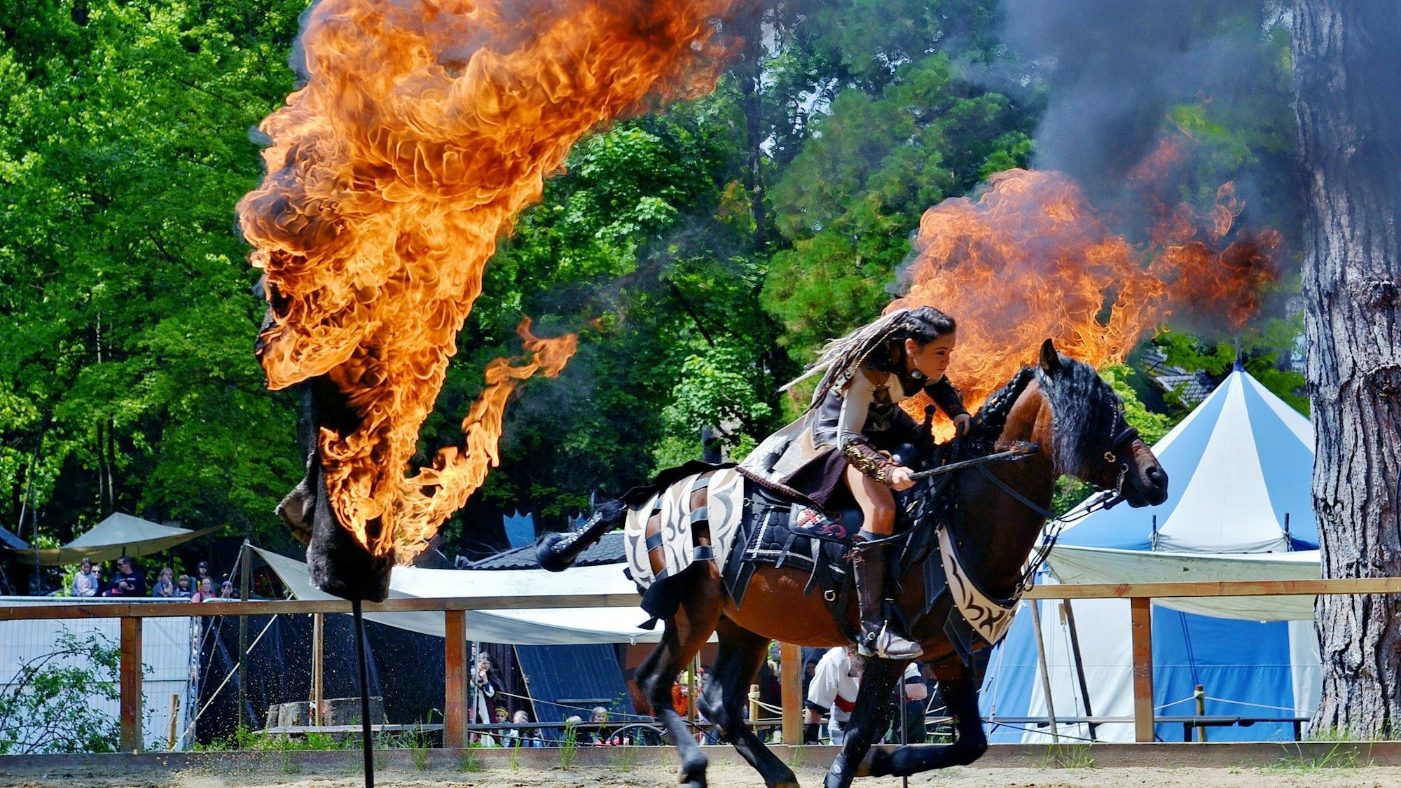 Eine Frau mit geflochtenen Haaren und Kampfausrüstung reitet zwischen zwei Feuertonnen hindurch.