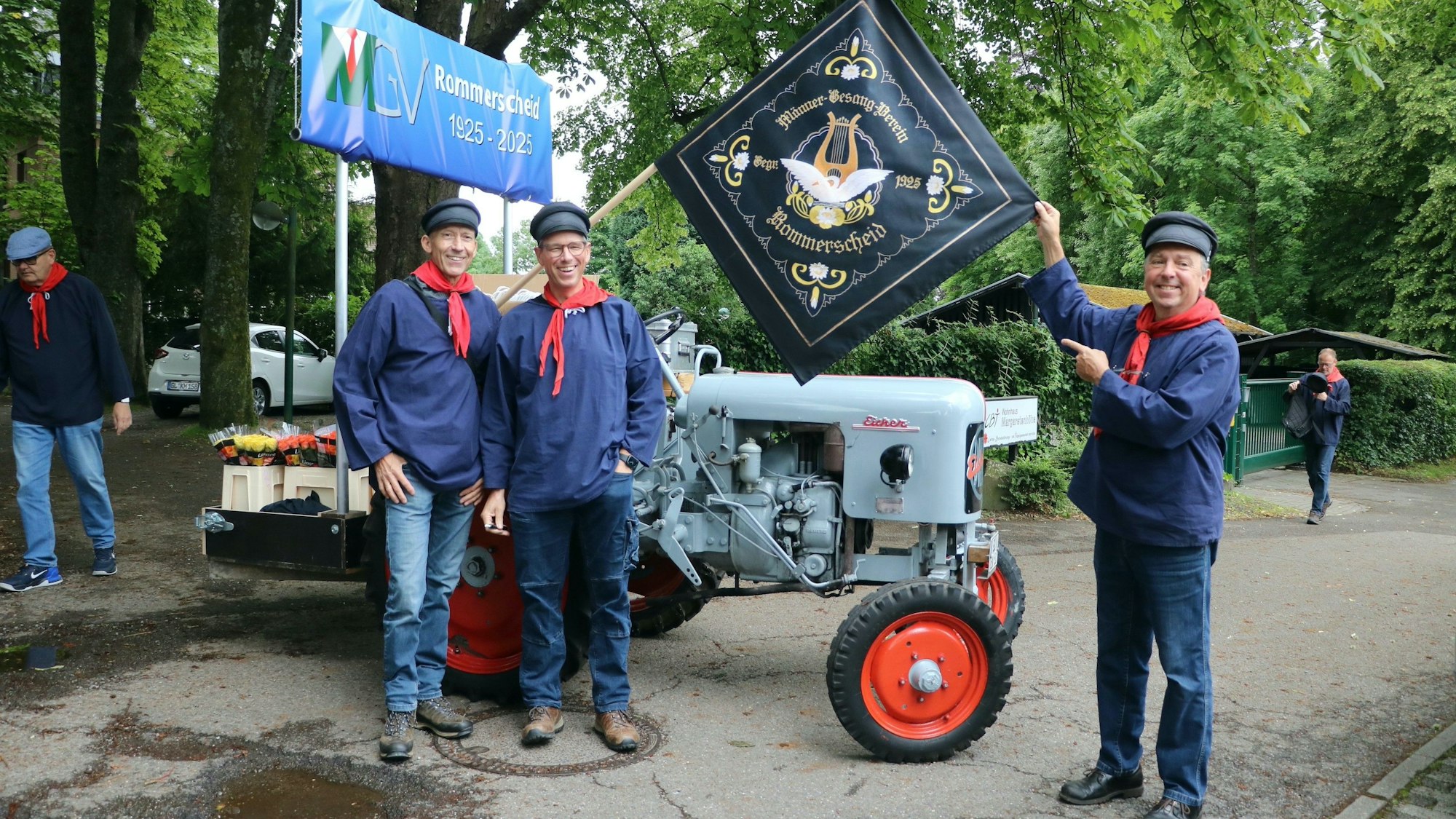 Drei Männer in bergischer blau-roter Tracht mit altem Traktor und Vereinsbanner.