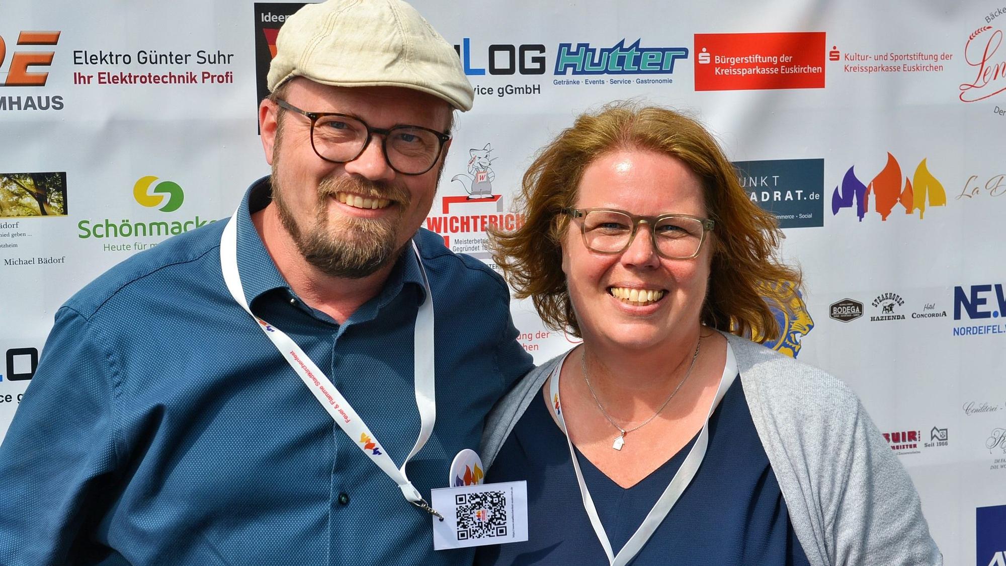 Eine Frau und ein Mann stehen vor einem Sponsorenbanner und lachen in die Kamera.