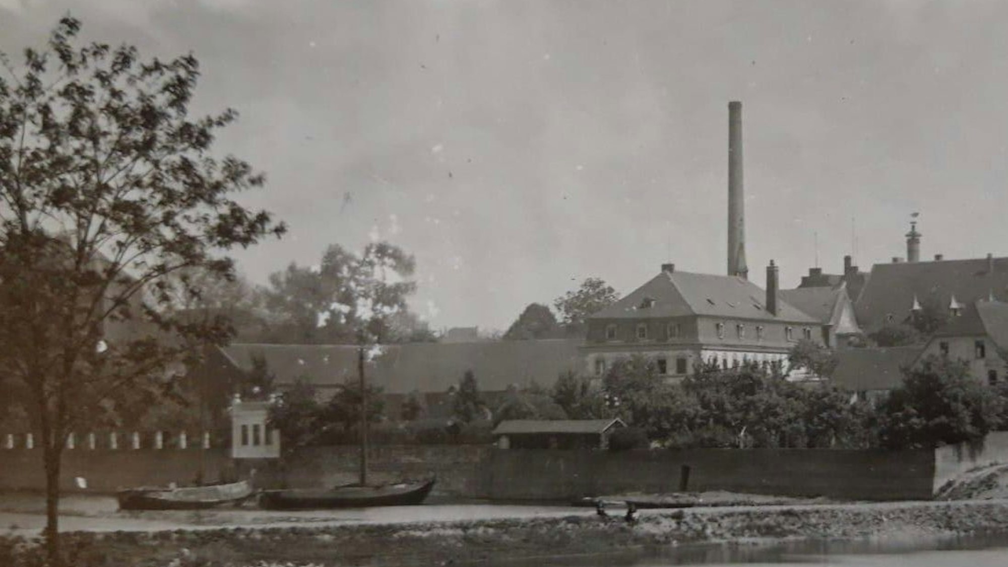 Ein Schwarz-Weiß-Bild zeigt eine Villa am Rhein mit einem kleinen Gebäude, das direkt am Ufer steht, Kähne liegen auf dem Wasser.