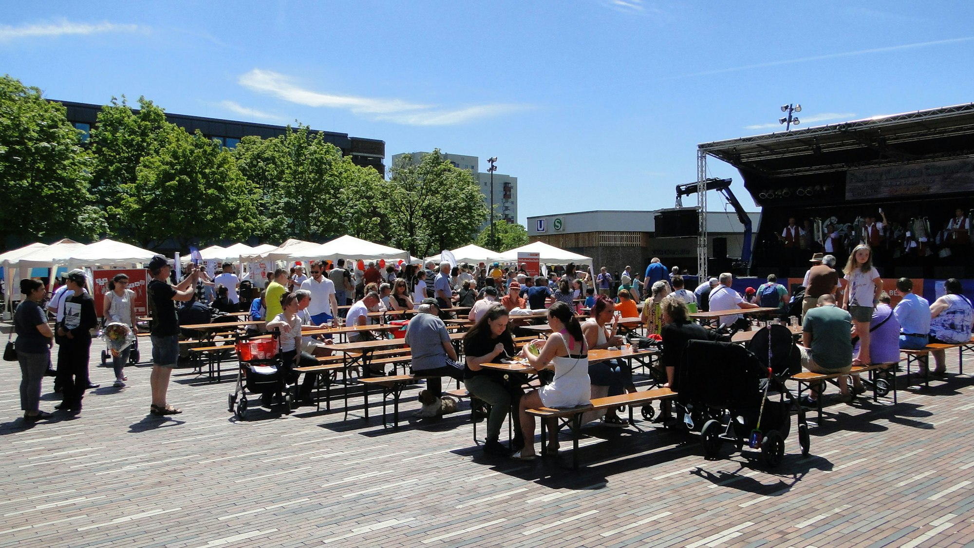 Zu sehen sind Menschen, die auf Biergarnituren sitzen, in der Sonne auf dem Pariser Platz in Chorweiler. Links im Bild sind weiße Pavillons und grüne Bäume zu sehen, recht eine Bühne.