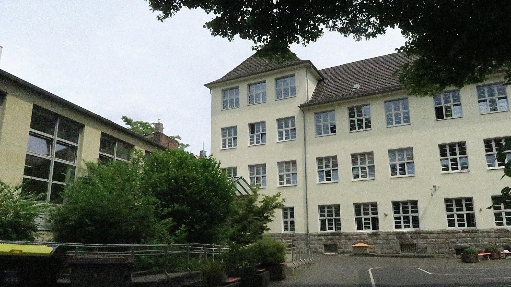 Ein mehrgeschossiges herkömmliches Schulgebäude mit vorgelagertem Schulhof und Grünanlage ist zu sehen.