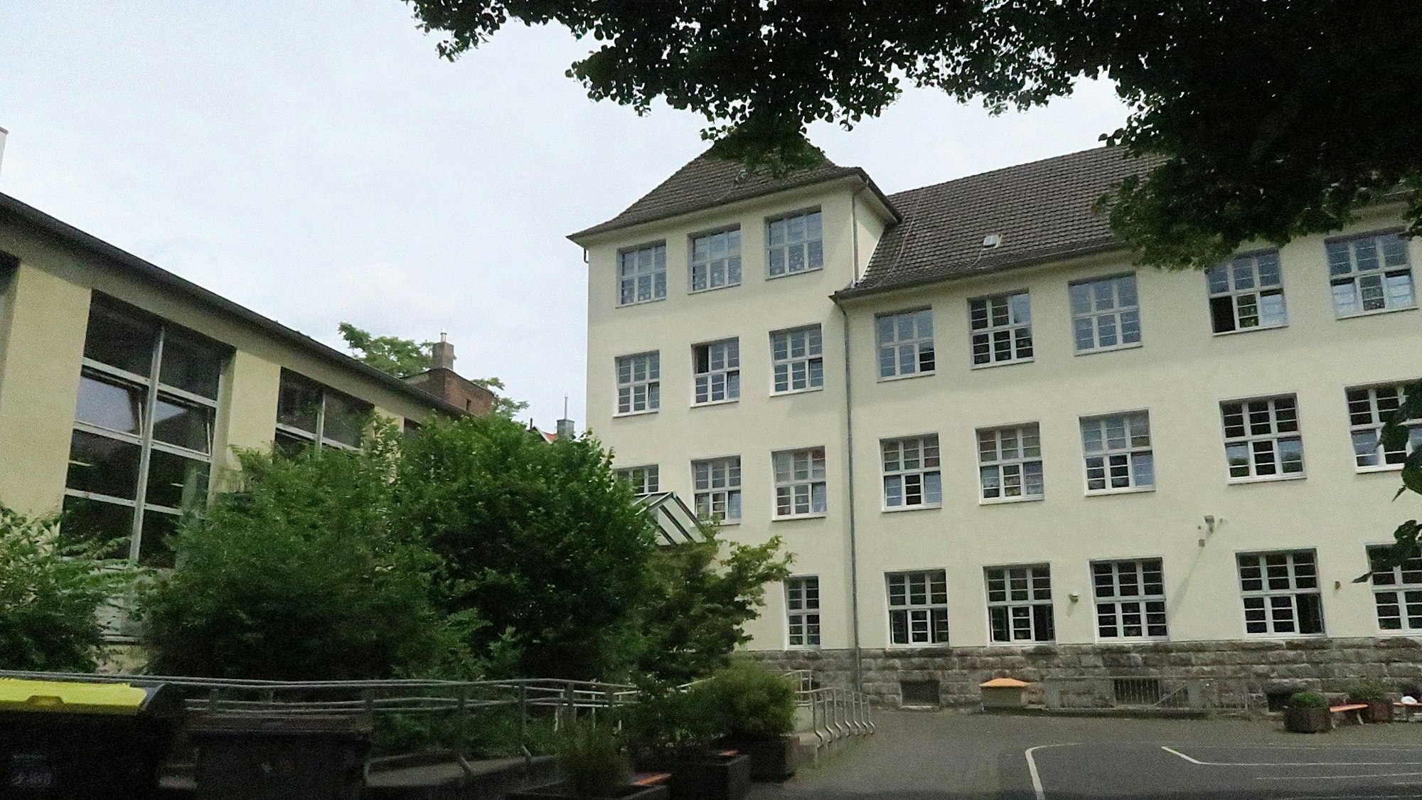 Ein mehrgeschossiges herkömmliches Schulgebäude mit vorgelagertem Schulhof und Grünanlage ist zu sehen.
