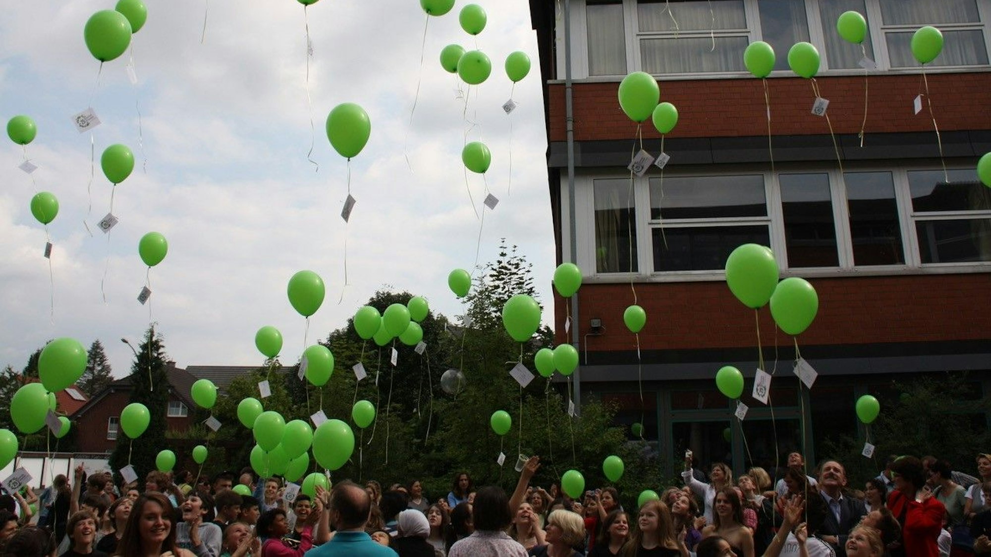 Viele Schüler und Eltern stehen auf dem Schulhof und lassen grüne Luftballons in den Himmel steigen.