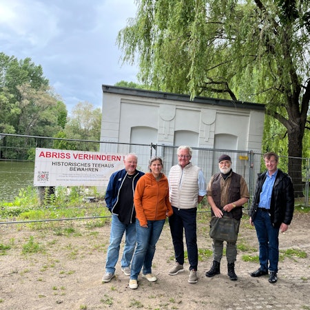 Vier Männer und eine Frau stehen vor einem kompakten alten Gebäude, das von einem Bauzaun abgesperrt ist.