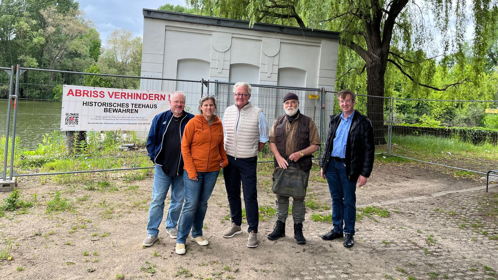 Fünf Menschen stehen vor einem kleinen Gebäude, das hinter einem Bauzaun steht.