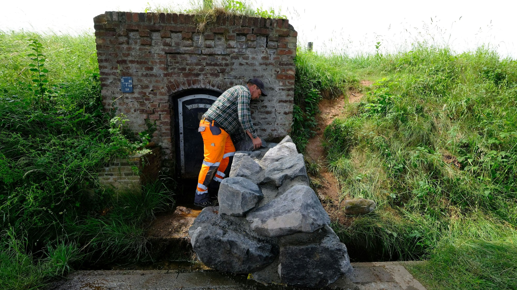 Ein Mitarbeiter des Bauhofs mit orangefarbener Arbeitshose und kariertem Hemd an der nun mit großen Natursteinblöcken verschlossenen Quelle in Lessenich.