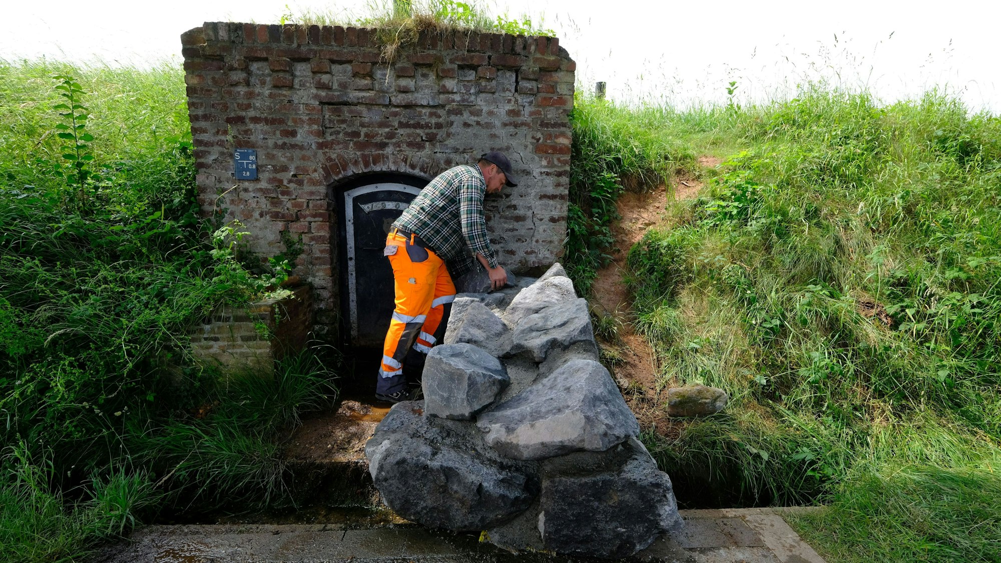 Ein Mitarbeiter des Bauhofs mit orangefarbener Arbeitshose und kariertem Hemd an der nun mit großen Natursteinblöcken verschlossenen Quelle in Lessenich.