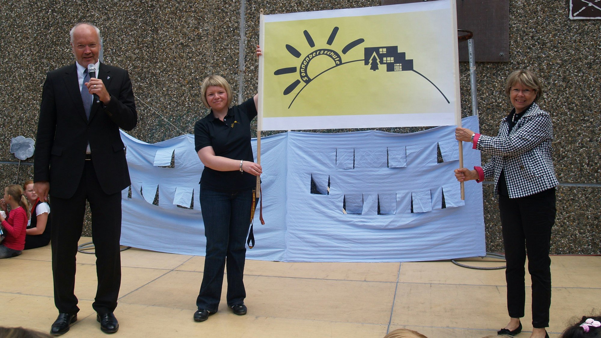 Zwei Frauen halten ein Schild mit einem gelb-schwarzen Logo hoch, das stilisiert eine Sonne und Gebäude auf einem Berg zeigt. Ein Mann links daneben hält ein Mikrofon.