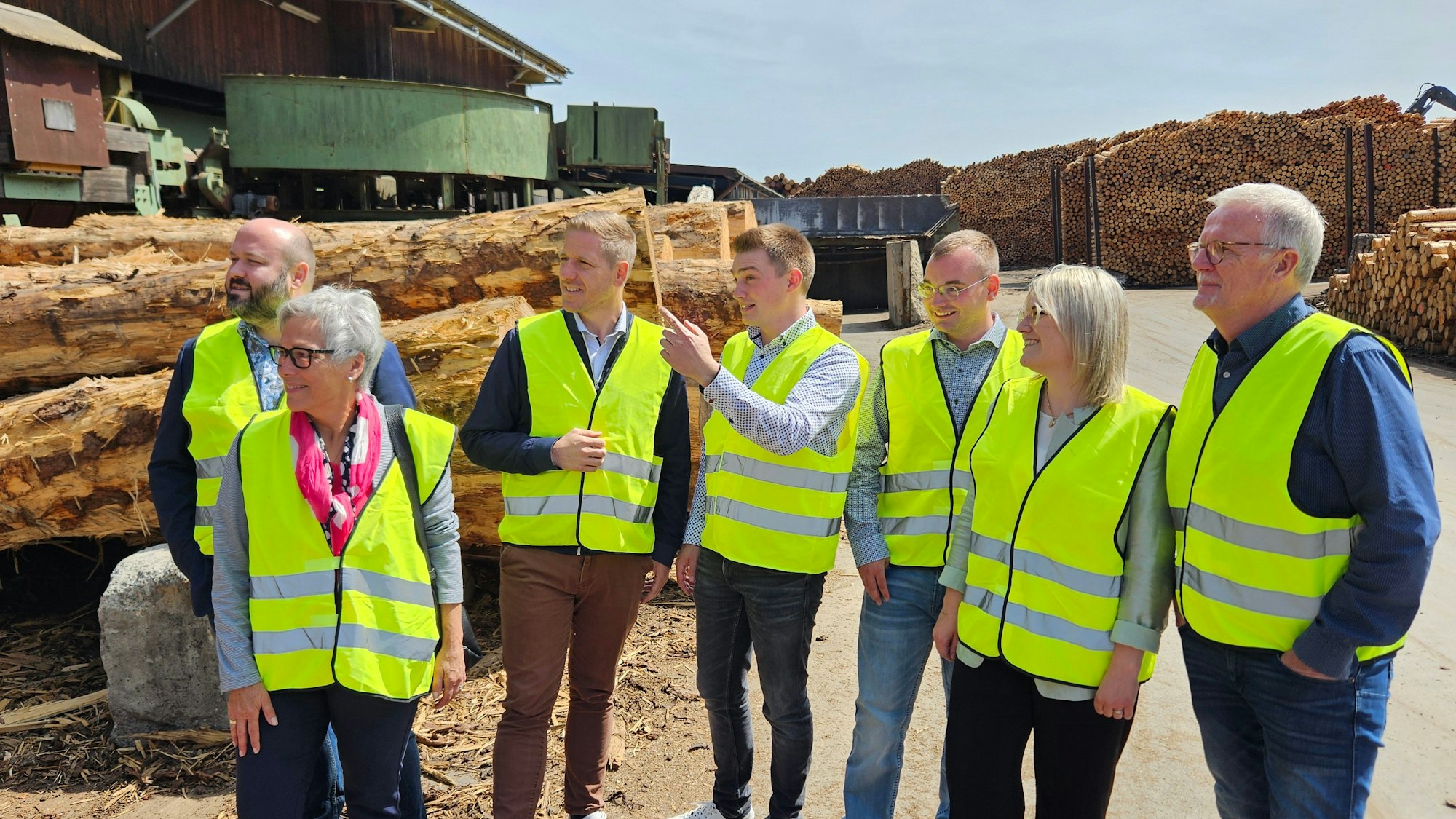Fünf Männer und zwei Frauen mit gelben Westen stehen nebeneinander, im Hintergrund sind große Stämme zu sehen.