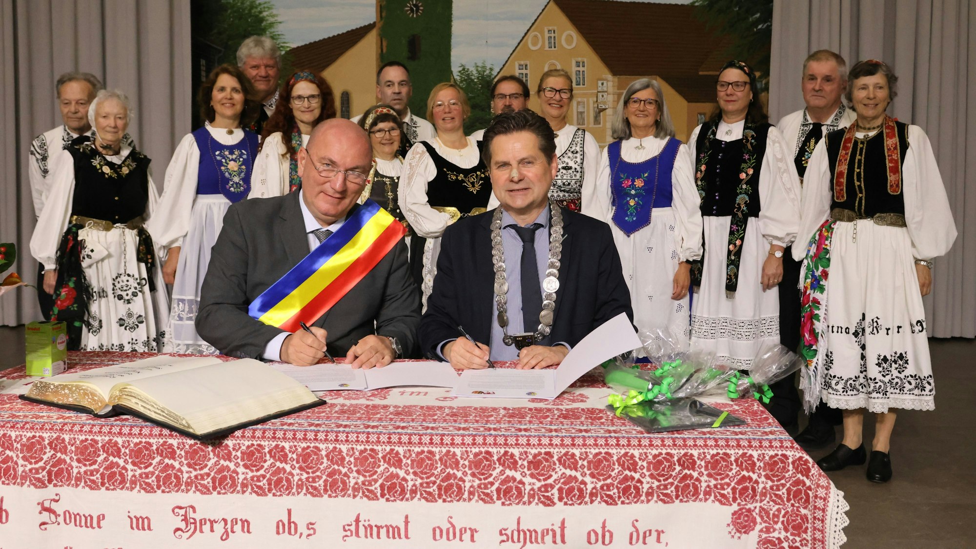 Zwei Männer an einem Tisch, dahinter eine Trachtengruppe.