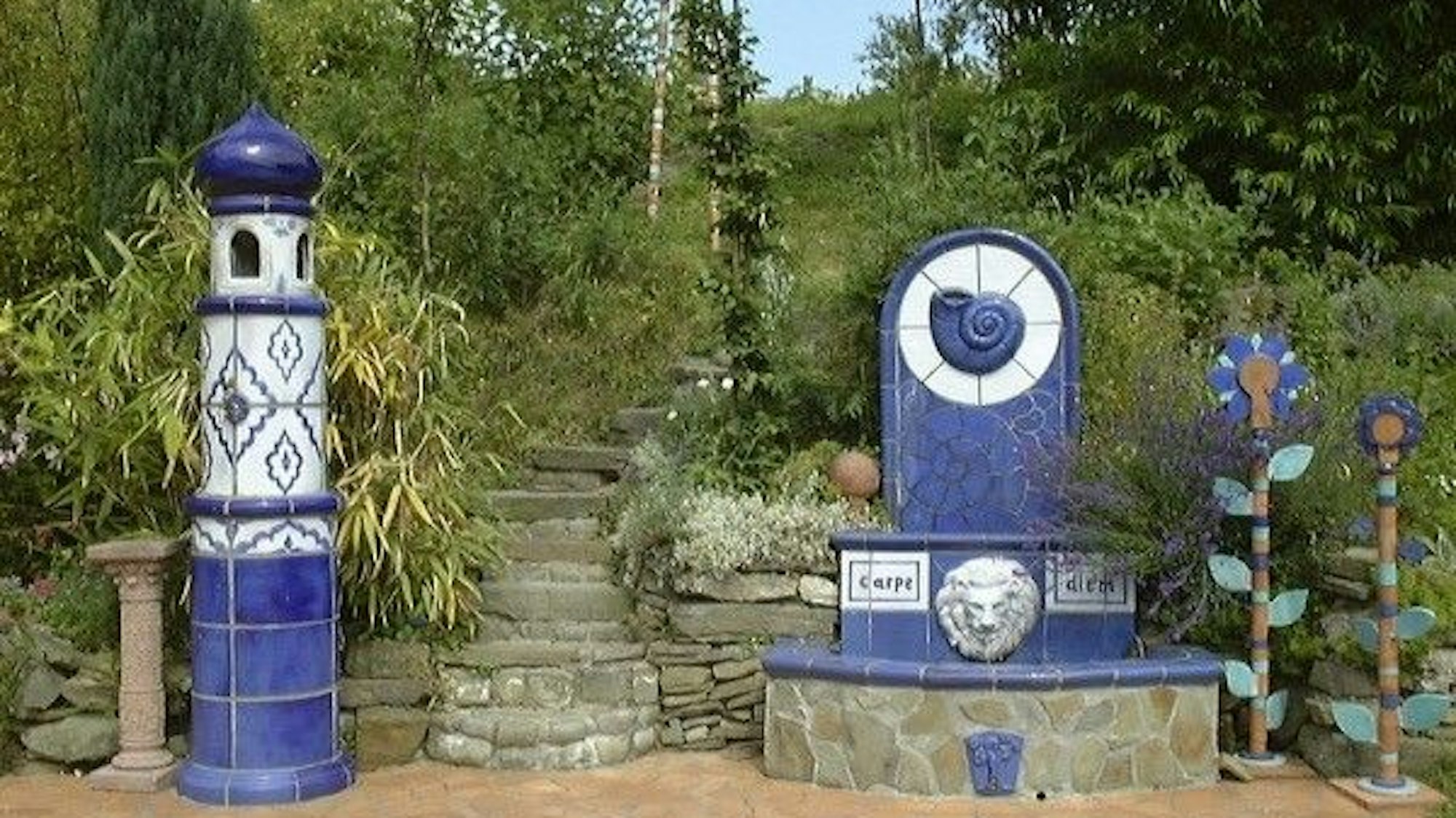 Ein blauer Brunnen und ein blauer Leuchtturm stehen vor einer Treppe, die ins Grüne führt.