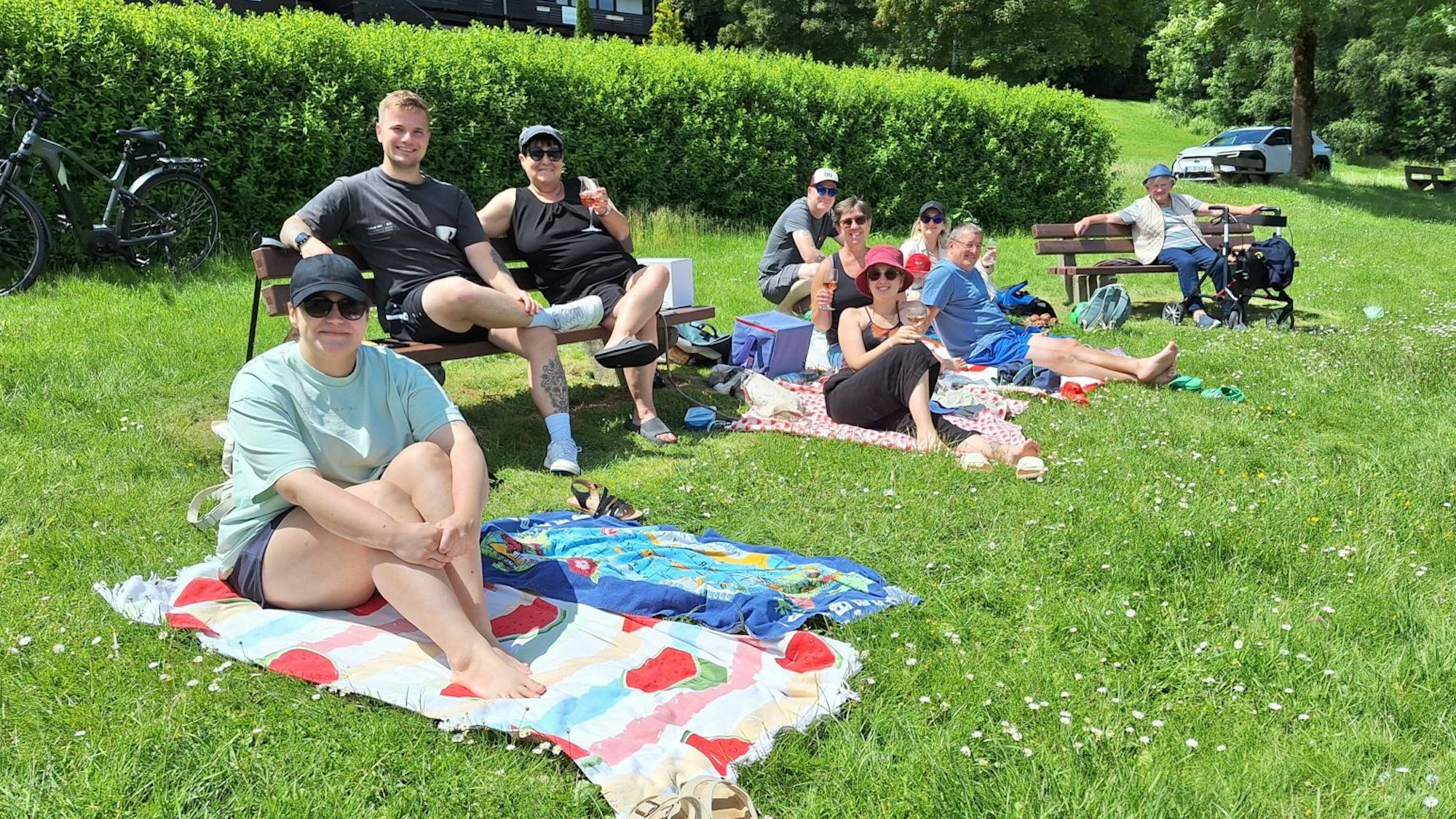 Das Bild zeigt Männer und Frauen, die sich auf einer Rasenfläche auf Sitzbänken und Decken niedergelassen haben.