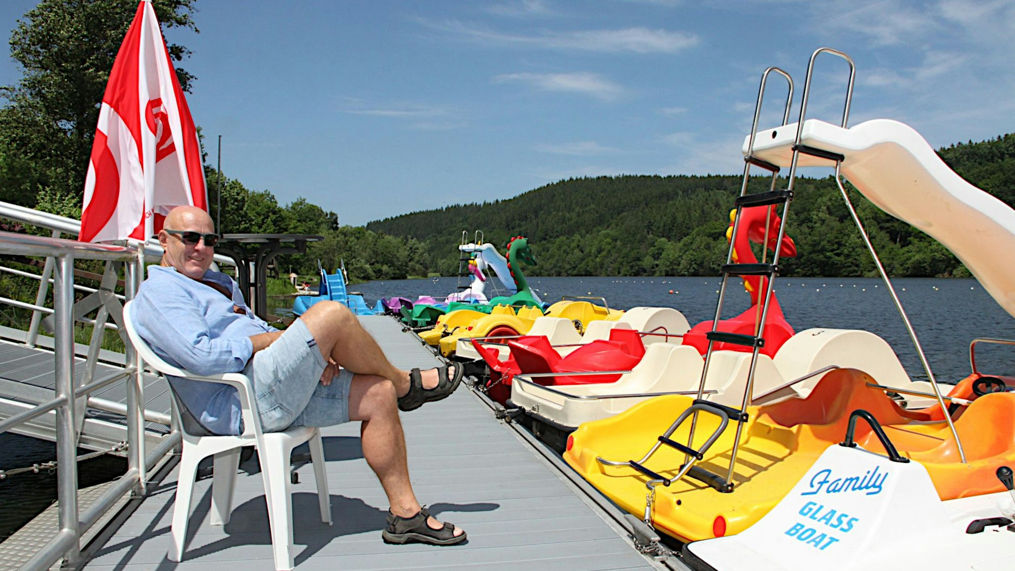 Das Bild zeigt einen Mann in Shorts auf einem Steg, an dem Tretboote in verschiedenen Farben liegen.