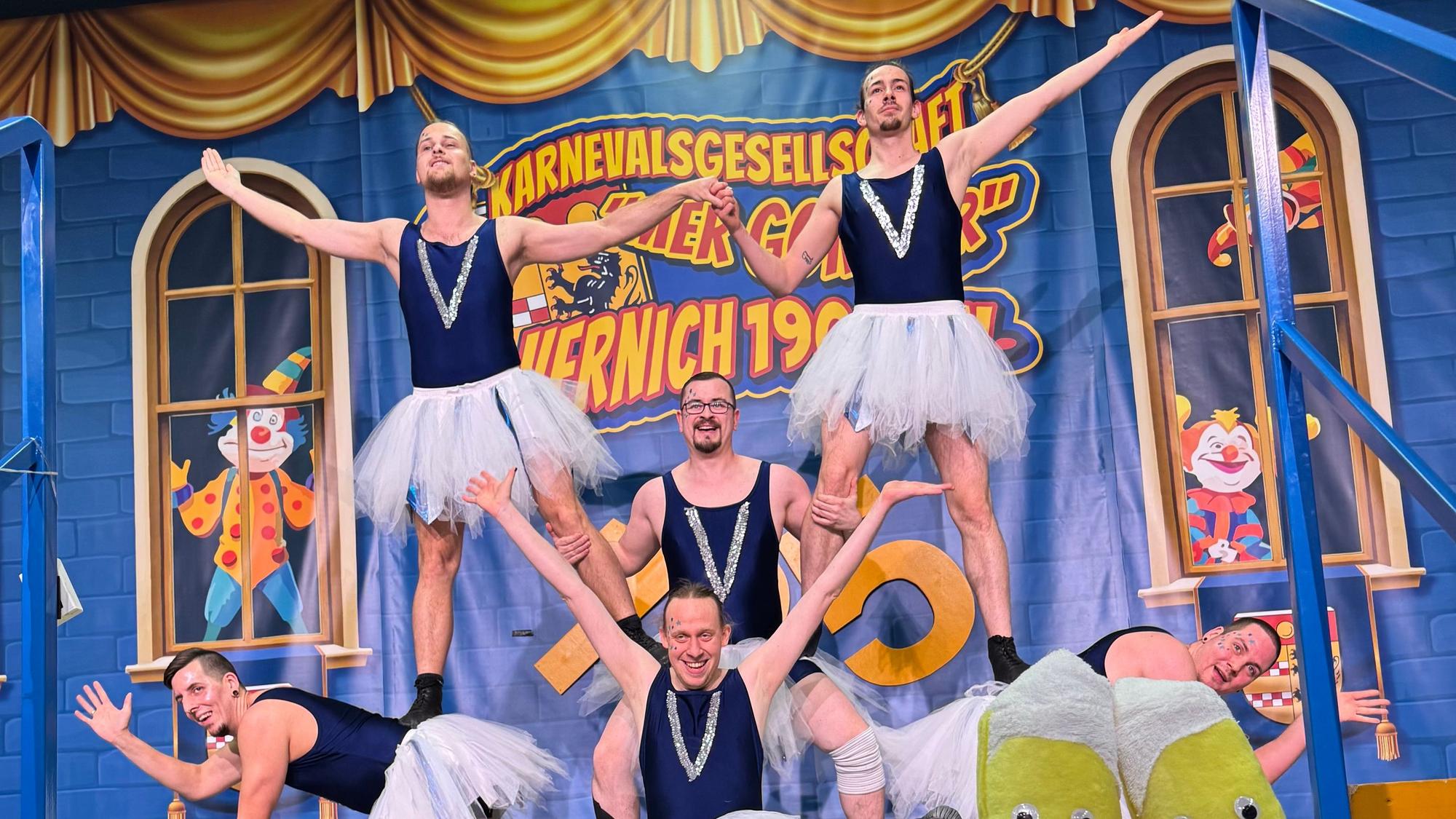 Sechs Männer in Tutus präsentieren auf einer Bühne eine Figur. Ihre Körper bilden mehrere Etagen.