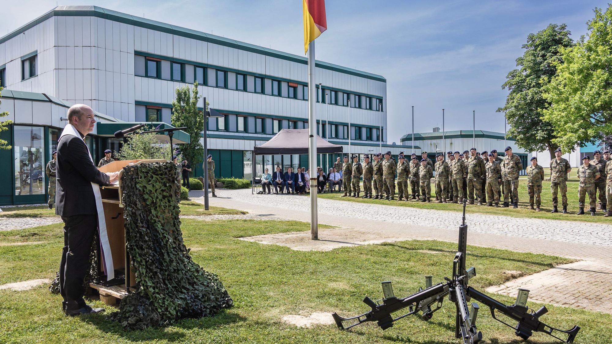 Vor dem Kasernengebäude spricht der katholische Militärpfarrer Lars Spohr zu den angetretenen Soldatinnen und Soldaten und Gästen. Vor ihm sind Sturmgewehre zusammengestellt.